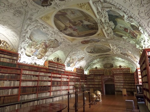 The monastery library is a long room with a low, arched ceiling that is profusely painted with human figures and scenes, presumably of religious subjects. The room’s walls are lined with bookshelves filled with antique-looking volumes. In the otherwise bare middle of the room are four large globes on stands and two small tables where people can sit or stand up to read.
Photographer: HutheMeow. Creative Commons Attribution-Share Alike 4.0 International license via Wikimedia Commons.